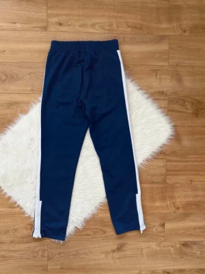 Blå Palm Angels mjukisbyxor med vita ränder - Mörkblå mjukisbyxor från Palm Angels med vita ränder längs sidorna och elastisk midja. Byxorna har raka ben och fickor framtill. Perfekta för en avslappnad och sportig stil.