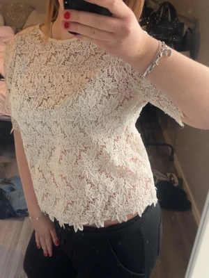 Vit spetsblus med blommönster - Superfin vit blus i spets med blommigt mönster och korta ärmar. Blusen är något croppad och har en rund halsringning. Perfekt att styla med både jeans och kjol för en trendig look.
