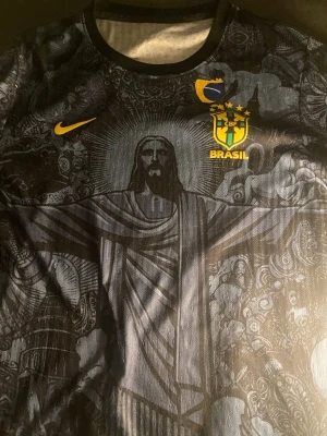 Svart Brasilien Nike fotbollströja - Säljer en svart fotbollströja från Nike med Brasilien CBF-emblem och gult Nike-logga. Tröjan har ett detaljerat motiv av Kristusstatyn och mönster över hela tyget. Perfekt för dig som vill sticka ut på planen.
