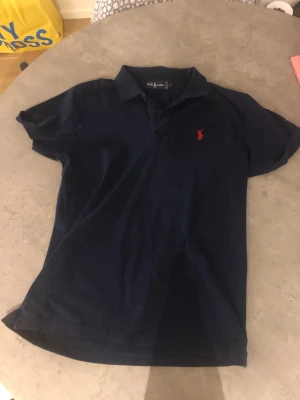 Marinblå pikétröja från Ralph Lauren - Klassisk marinblå pikétröja från Ralph Lauren med korta ärmar och röd broderad logga på bröstet. Tröjan har krage och knäppning framtill. Perfekt för en stilren och sportig look. Storleken är XXL men sitter mer som en M/L (OBS PRIS GÅR ATT DISKUTERA!)