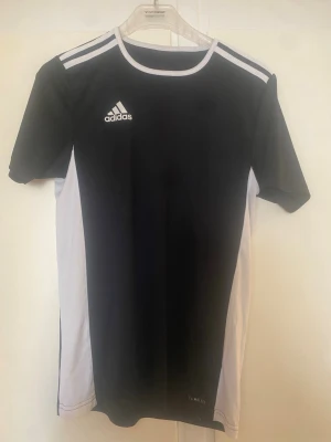 Svart och vit Adidas sporttröja - Snygg svart och vit sporttröja från Adidas med klassiska vita ränder på axlarna och logga på bröstet. Tillverkad i lätt och ventilerande material, perfekt för träning eller fotboll. Rund halsringning och korta ärmar ger en sportig look.