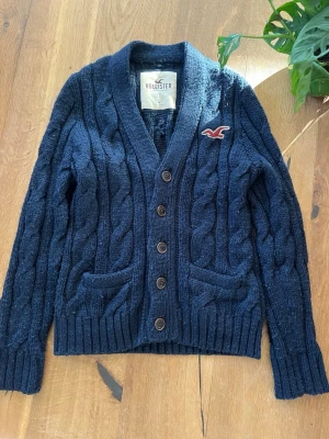 Mörkblå kabelstickad kofta Hollister - Mörkblå kabelstickad kofta från Hollister med fyra knappar framtill, två fickor och ribbade muddar. Koftan har en klassisk v-ringad krage och Hollisters logga broderad på bröstet. Perfekt för lager-på-lager och chill stil.