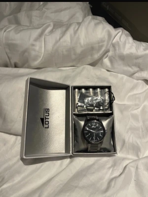 Svart klocka från Lotus i metall - Stilren svart Lotus-klocka med metallarmband och klassisk rund urtavla. Svart urtavla med vita detaljer och datumvisning. Levereras i originalask med extra länkar. Perfekt för dig som gillar en clean och modern stil. Klockan har några synliga skraprepor (se bild). Nypris ca 1400 kr. Pris kan diskuteras vid snabb affär. Ps den har några repor men ingen stor skillnad.