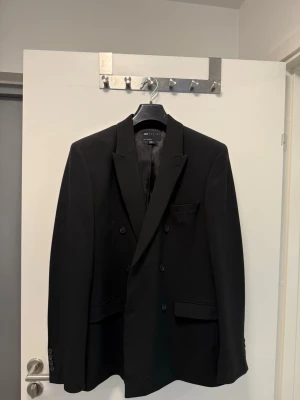 Svart dubbelknäppt kavaj ASOS DESIGN - Stilren svart kavaj från ASOS DESIGN med dubbelknäppning och klassisk passform. Kavajen har smala slag, tre framfickor och knappar vid ärmslut. Perfekt för en dressad look och tillverkad i ett slätt tyg med fodrad insida.