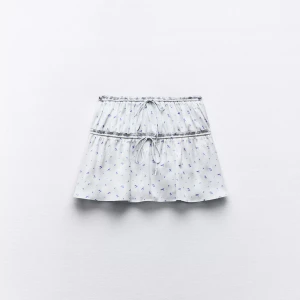 Vit blommig kortkjol med snörning - Supersöt vit kortkjol med små blå blommor och dubbla snörningar i midjan och på höften. Kjolen har en lätt volangig form och är perfekt för en fräsch och somrig look. Tunt och luftigt material som känns skönt mot huden. 