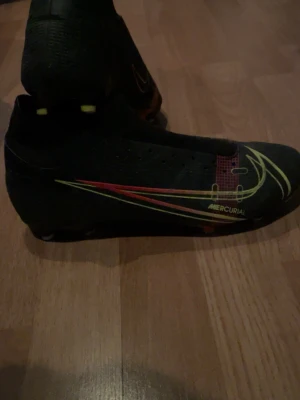 Nike Mercurial svarta fotbollsskor - Säljer ett par svarta Nike Mercurial fotbollsskor med coola gula och röda detaljer. Skorna har en strömlinjeformad design och mönstrad sula med fasta dobbar för bra grepp på planen. Perfekta för dig som vill sticka ut på matchen! Tvättar såklart skorna in dom skickas 