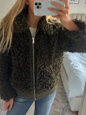 Mörkgrön teddyjacka med dragkedja - Säljer en fluffig mörkgrön teddyjacka med krage och grov dragkedja framtill. Jackan har en boxy passform och muddar vid ärmslut och nederkant. Perfekt för dig som vill ha en mysig och trendig look i höst och vinter.
