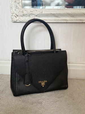 Svart handväska från Prada  - Snygg svart handväska från Prada  med guldfärgade detaljer och dubbla handtag. Väskan har ett elegant triangulärt lock, dragkedja upptill och en dekorativ nyckelring i metall. Perfekt för dig som gillar stilrena accessoarer.
