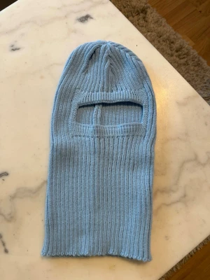 Ljusblå ribbstickad balaclava - Säljer en ljusblå ribbstickad balaclava i mjukt material. Mössan har en öppning för ögonen och näsan, perfekt för kalla dagar. Enkel och stilren design utan synligt märke, passar dig som vill ha något både praktiskt och snyggt.