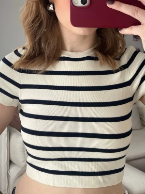 Randig croppad topp i ribbad bomull - Säljer en croppad topp med svarta och vita horisontella ränder. T-shirten har korta ärmar, rund halsringning och är gjord i ett ribbat bomullsmaterial som sitter tajt. Perfekt till höga jeans eller kjol för en trendig look.