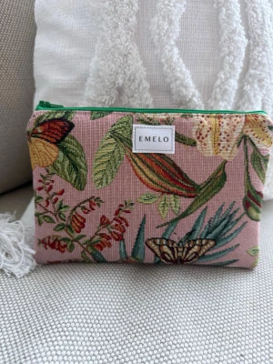 Blommig necessär från Emelo - Säljer en snygg necessär från Emelo med broderat blommönster i grönt, rött och gult på en rosa bakgrund. Väskan har en grön dragkedja och är tillverkad i ett tygmaterial med struktur. Perfekt för smink eller småprylar.