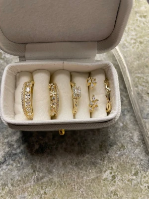 Fem  ringar guld pläterade  med guld 18k med stenar och blommor - Säljer ett set med fem guld pläterade ringar i guld 18k i olika designer. Ringarna har gnistrande stenar och detaljer som blommor, löv och rektangulär sten. Perfekta för att mixa och matcha för en trendig look. Ringarna är justerbara i storleken förutom den första ringen från höger som har ett mått på 1,6 cm diameter 