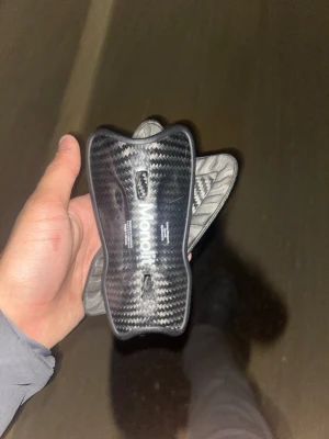 Monolit benskydd i kolfiber - Säljer ett par svarta benskydd från Monolit i kolfiber med snygg, glansig finish och ergonomisk form. Nästan helt oanvända. Perfekta för fotbollsträning eller match när du vill ha lätt och starkt skydd. Diskret design med vit logga på framsidan.