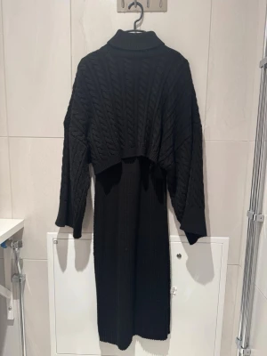 Svart stickad långklänning med tröja i polokrage - Svart långklänning i stickat material med polokrage och grovt kabelstickat mönster upptill. Klänningen har långa ärmar och en loose passform upptill, vilket ger en cool och avslappnad look. Perfekt för dig som gillar mysiga och trendiga plagg.