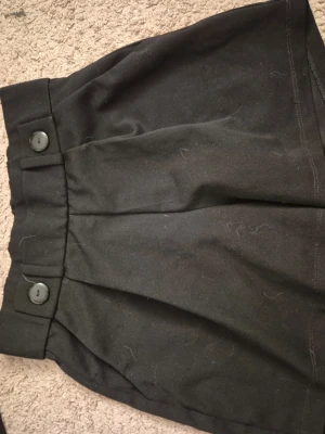 Svarta högmidjade shorts med knappar - Snygga svarta högmidjade shorts med två stora dekorativa knappar framtill och lätt rynkad midja. Shortsen har en loose fit och är tillverkade i ett mjukt tyg, perfekta för en chill och stilren look.