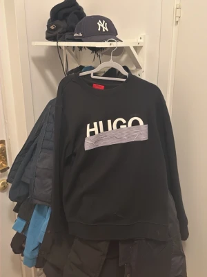 Svart sweatshirt från HUGO - Snygg svart sweatshirt från HUGO med vit logga och grafiskt tryck över bröstet. Tröjan har rund halsringning och avslappnad passform, perfekt för en casual look. Tillverkad i mjukt material för extra komfort.