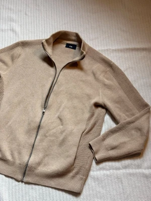 Beige ziptröja från C&A - Beige stickad ziptröja från C&A med hög krage och hel dragkedja framtill. Tröjan har ribbade muddar och detaljer längs ärmarna samt en enkel, stilren design som passar till många olika outfits. Perfekt för lager på lager och en avslappnad look.