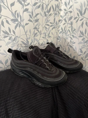Svarta Nike Air Max 97 sneakers - Säljer ett par svarta Nike Air Max 97 sneakers med ikoniska vågformade linjer och reflekterande detaljer. Skorna har en låg siluett, synlig Air Max-sula och är tillverkade i mesh och syntetmaterial. Perfekta för dig som gillar streetwear och sportig stil.  Sko snöret på vänster sko saknar plast toppen se på sista bilden 