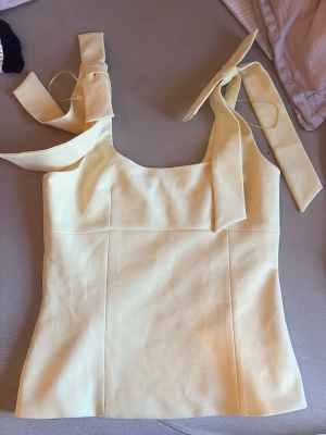 Gul linnetopp med knyt från Zara - Supersöt ljusgul linnetopp från Zara med breda axelband som knyts i rosetter. Toppen har en rak passform och är tillverkad i ett mjukt material som känns skönt mot huden. Perfekt för en fräsch och trendig look.