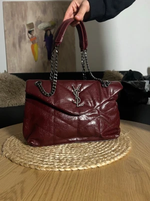 Vinröd YSL handväska i läder - Snygg vinröd handväska från YSL i quiltat läder med silverfärgad kedja och ikonisk YSL-logga framtill. Väskan har både handtag och axelrem i kedja, perfekt för att lyfta vilken outfit som helst.