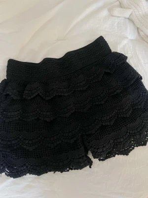 Svarta virkade shorts med volang - Snygga svarta shorts med virkade lager och volangdetaljer. De har en bred, elastisk midja och är perfekta för en trendig och avslappnad look. Materialet är mjukt och luftigt, vilket gör dem sköna att bära under varma dagar.