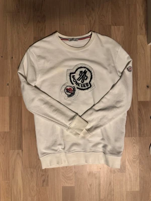 Vit stickad tröja från Monclerr - Snygg vit stickad tröja från Monclerr med stort broderat logomärke på bröstet och en mindre logga på ärmen. Tröjan har rund halsringning och långa ärmar. Perfekt för dig som gillar stilrena och exklusiva plagg.