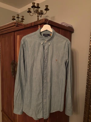 Ljusblå skjorta från Polo Ralph Lauren - Klassisk ljusblå skjorta från Polo Ralph Lauren med button-down krage och broderad logga på bröstet. Skjortan har lång ärm och knäppning framtill. Perfekt för en stilren och avslappnad look. Storlek L slimfit, sitter true to size!