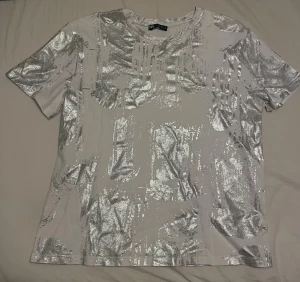 Vit t-shirt med silvermönster - Säljer en vit t-shirt från Zara med unikt silvermönster över hela plagget. T-shirten har korta ärmar och en klassisk rund halsringning. Materialet känns mjukt och skönt, perfekt för dig som vill sticka ut lite extra.