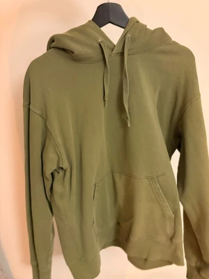 Olivgrön hoodie med magficka - Snygg olivgrön hoodie med huva och dragsnören. Klassisk modell med lång ärm och stor magficka framtill. Perfekt för en avslappnad streetstyle-look. Mjuk och skön bomullskänsla. Från Paul&Friends.