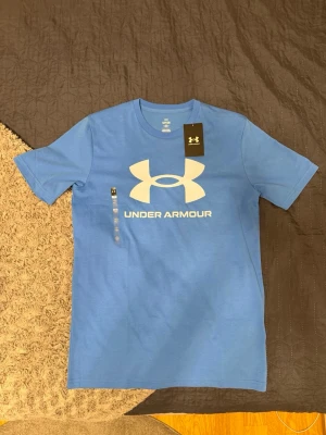Sprillansny Under Armour t-shirt  - Spillansny snygg blå t-shirt från Under Armour med stor vit logga och text på bröstet. Klassisk passform med korta ärmar och rund halsringning. Perfekt för en sportig och avslappnad stil. Vid funderingar eller likanande är det bara att höra av sig! 