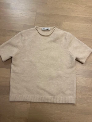 Beige stickad t-shirt från Zara - En stilren beige stickad t-shirt från Zara med korta ärmar och rund halsringning. Tröjan har ribbade muddar vid ärmslut och nederkant, vilket ger en clean och modern look. Aldrig använd så som ny! Passar xs-m