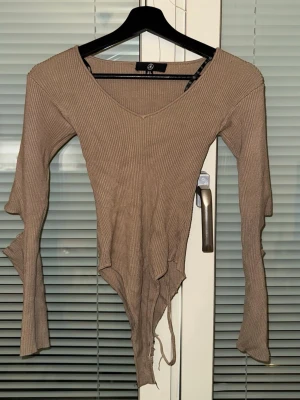Beige ribbad body med V-ringning - Trendig beige body från SHEIN med ribbad struktur och V-ringad hals. Långa ärmar med coola slitsdetaljer och asymmetrisk nederkant. Perfekt för att skapa en edgy look. Mjukt och stretchigt material som sitter snyggt på kroppen.