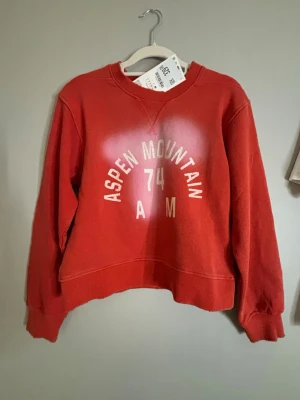 Röd zara sweatshirt ny med pris lapp - Röd sweatshirt med blekta fläckar och vit text framtill och baktill. På framsidan står det 'Aspen Mountain 74 AM' och på baksidan 'Aspen Mountain 1974 Rugby U.S.A Western Colorado'. Tröjan har rund hals, långa ärmar och avslappnad passform. Materialet ser ut att vara mjuk bomullsmix.