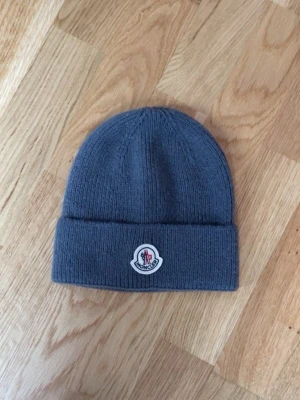 Grå Moncler mössa med logga - Snygg grå ribbstickad mössa från Moncler med uppvikt kant och klassisk Moncler-logga framtill. Perfekt accessoar för kyliga dagar och enkel att matcha med olika outfits. Materialet är mjukt och bekvämt.