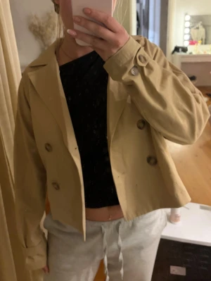Kort beige trenchcoat med knappar - Stilren kort trenchcoat i beige med dubbelknäppning och bred krage. Jackan har dekorativa knappar på ärmarna och en avslappnad passform. Perfekt för dig som vill ha en trendig och enkel look. Materialet känns som bomull eller en bomullsblandning.