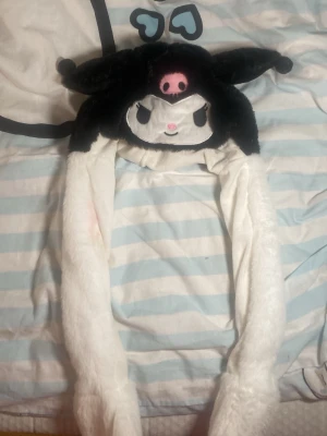Kuromi mössa med långa öron - Söt och mjuk mössa i form av Kuromi från Sanrio. Hatten är svart och vit med broderat ansikte, rosa detaljer och långa öron som hänger ner. Tillverkad i plyschmaterial, perfekt för dig som gillar kawaii-stil och vill sticka ut. Har fläckar från smink. Kan lyfta på öronen och lyser med färger. Har tre olika varianter av blickningar