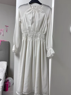 Vit långärmad klänning - Vit långklänning med markerad midja och långa ärmar med lätt volang vid ärmslut. Klänningen har en rund halsringning och är tillverkad i ett tunt, lätt material som ger ett luftigt intryck.