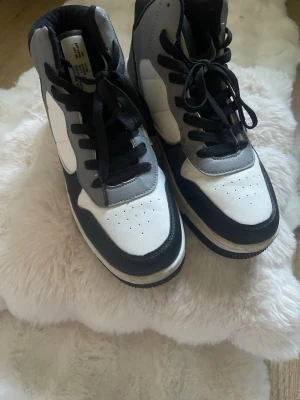 Svarta och vita high top sneakers - Snygga high top sneakers i svart, vitt och grått med klassisk siluett och svarta skosnören. Ovandelen är i syntetmaterial med perforerade detaljer på tån för extra stil. Perfekta för dig som gillar streetwear och vill ha en clean look.