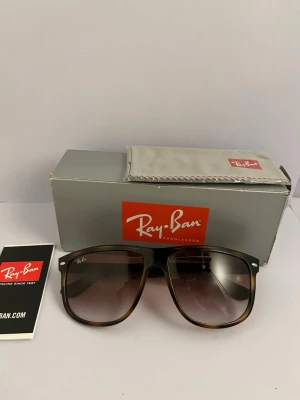 Rayban Boyfriend - Snygga solglasögon från Ray-Ban med brunmelerad båge och stora, fyrkantiga glas. Glasen är tonade i brunt och bågen har en klassisk design med diskret logga på linsen. Perfekta för att lyfta din stil under soliga dagar.