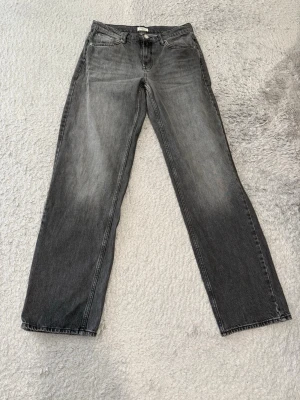 Grå raka jeans med tvättad look - Säljer ett par grå jeans med rak passform och snyggt tvättad finish. Jeansen har klassiska fem fickor, bälteshällor och dragkedjegylf. Materialet är denim i bomull och färgen är en cool grå nyans med slitningar framtill.