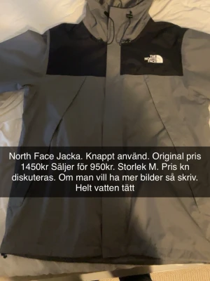 Grå regnjacka från The North Face - Snygg grå regnjacka från The North Face med svarta axelpartier och huva. Jackan är helt vattentät och har dragkedja framtill. Perfekt för regniga dagar och outdoor-äventyr. Klassisk logga på bröstet och stilren design.