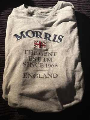 Grå tjoktröja från Morris - Grå sweatshirt från Morris med broderad text och brittisk flagga på bröstet. Tröjan har rund halsringning, långa ärmar och ribbade muddar. Perfekt för en avslappnad och stilren look.