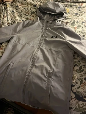 Grå vindjacka från Under Armour - Snygg grå vindjacka från Under Armour med huva och dragkedja framtill. Jackan har två fickor med dragkedja och en diskret logga på bröstet. Perfekt för blåsiga dagar och sportiga outfits.