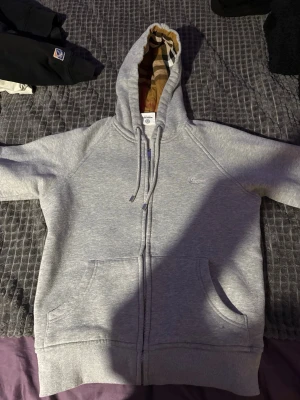 Grå zip-hoodie från Burberry - Snygg grå hoodie från Burberry med dragkedja och klassiskt rutigt foder i huvan. Mjuk bomullsblandning, två fickor fram och diskret Burberry-logga på bröstet. Perfekt för en chill och stilren look.