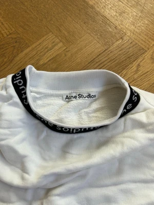 Vit sweatshirt från Acne Studios - Vit långärmad sweatshirt från Acne Studios med svart ribbad krage där varumärkets namn är invävt. Tröjan har en avslappnad passform och breda muddar vid ärmslut och nederkant. Perfekt för en clean och stilren look. Storleken är M men tröjan är väldigt loose/oversized i modellen.