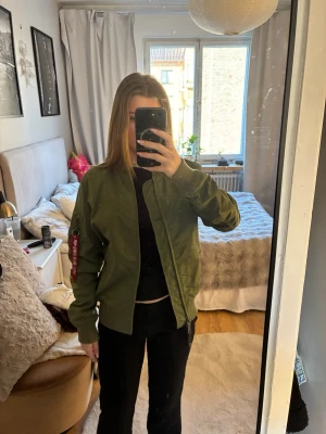 Grön bomberjacka från Alpha Industries - Säljer en klassisk grön bomberjacka från Alpha Industries med ribbade muddar och dragkedja framtill. Jackan har en röd tag på ärmen och flera fickor. Perfekt för dig som vill ha en cool och tidlös look. Passar S och XS