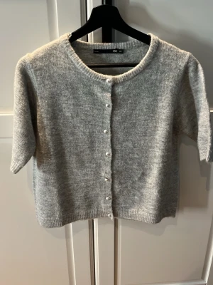 Grå kofta från 157 med pärlknappar - Mysig grå kofta från 157 med rund halsringning och pärlknappar framtill. Koftan har korta ärmar och är stickad i ett mjukt material som passar perfekt till lager-på-lager-stil. Enkel och stilren design som funkar till många outfits.