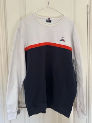 Le Coq Sportif sweatshirt blå/vit/röd - Snygg sweatshirt från Le Coq Sportif i färgblockad design med vit överdel, marinblå nederdel och röd rand över bröstet. Broderad logga på vänster bröst. Mjuk bomullskvalitet och avslappnad passform, perfekt för en sportig och casual look.
