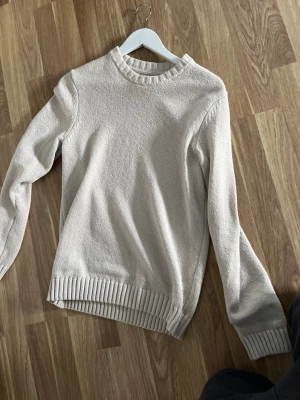 Beige stickad tröja med rund hals från Brothers - En stilren beige stickad tröja med rund halsringning och ribbade muddar vid ärmslut, hals och nederkant. Tröjan har en klassisk passform och är perfekt för lager-på-lager. Materialet ser mjukt och bekvämt ut, perfekt för kyligare dagar.