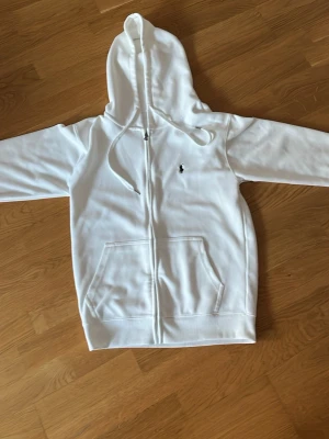 Vit hoodie från Polo Ralph Lauren - Snygg vit hoodie från Polo Ralph Lauren med dragkedja framtill, huva med dragsko och klassisk broderad logga på bröstet. Mjuk bomullskvalitet och känguruficka gör den både stilren och bekväm. Perfekt för en avslappnad och trendig look.